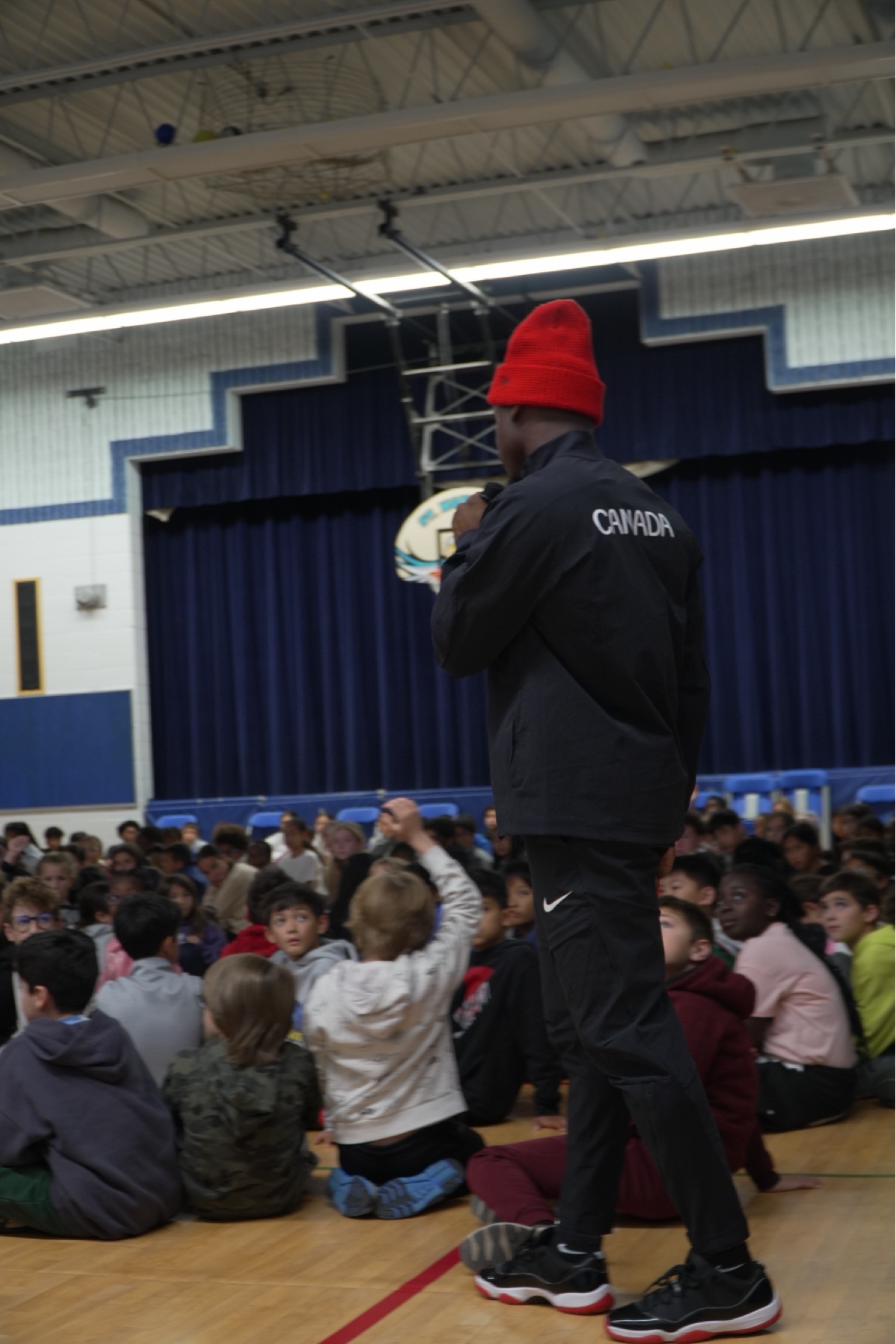 Duan Asemota in a Team Canada jacket speaking to elementary school students sitting on a gym floor